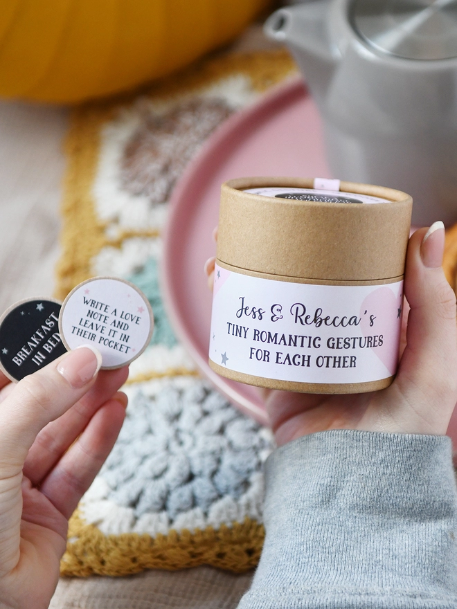 A cardboard jar with a pale pink label that reads "Tiny romantic gestures for each other" is being held in one hand. Two wooden tokens are held in another hand.