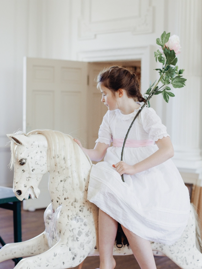 A girl sits on a rocking horse in a while dress with a pink sash holding a flower
