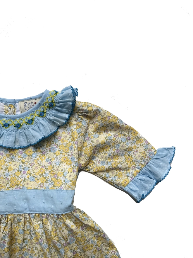 Detail of A yellow floral dress with a blue swiss dot frill collar and sleeves and a blue sash. Smocked detailing around the neckline.