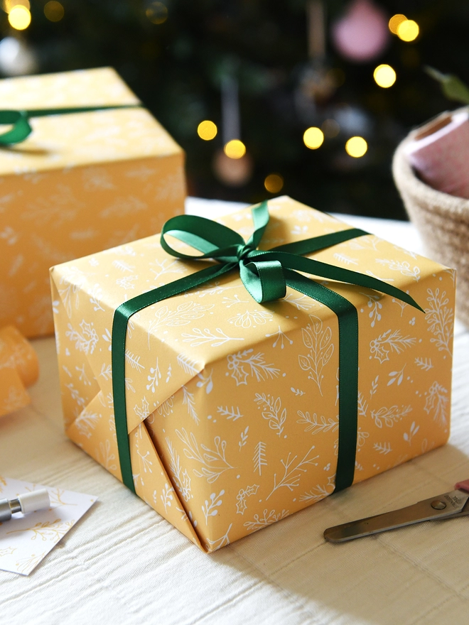 A gift wrapped in yellow botanical wrapping paper with green ribbon is on a table in front of a Christmas tree.