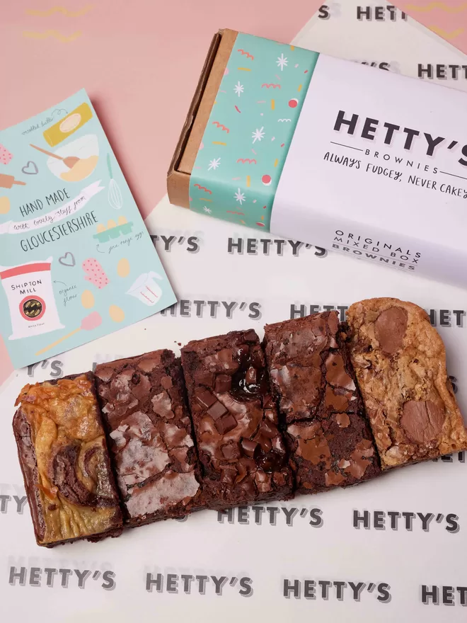 Five slices of Hetty's brownie original flavours of marbled cheesecake, salted caramel, classic fudge, cookie dough and chocolate orange in a flat lay with branded box and postcard