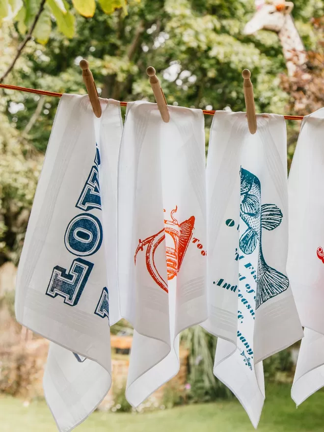 A selection of Mr.PS printed cotton hanndkerchiefs pegged on a washing line in a pretty garden.
