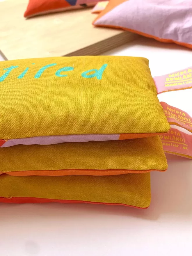 Stack of lavender eye pillows in yellow fabric with neon turquoise letters which read 'so tired' 