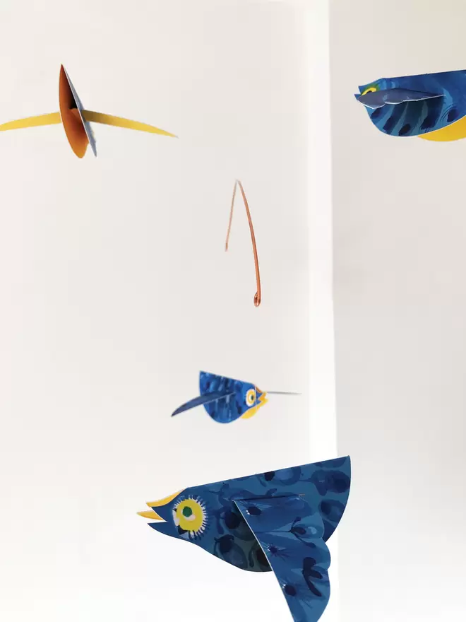 Four bright blue patterned paper birds mobile hanging against a white wall. They show sunshine-yellow bellies.