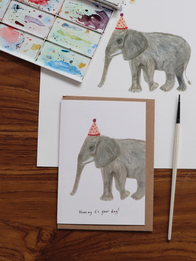  Desk Shot Of The Elephant Greeting Card Sitting Along Side The Original Hand Painted Watercolour Illustration, Paintbrush and Palette