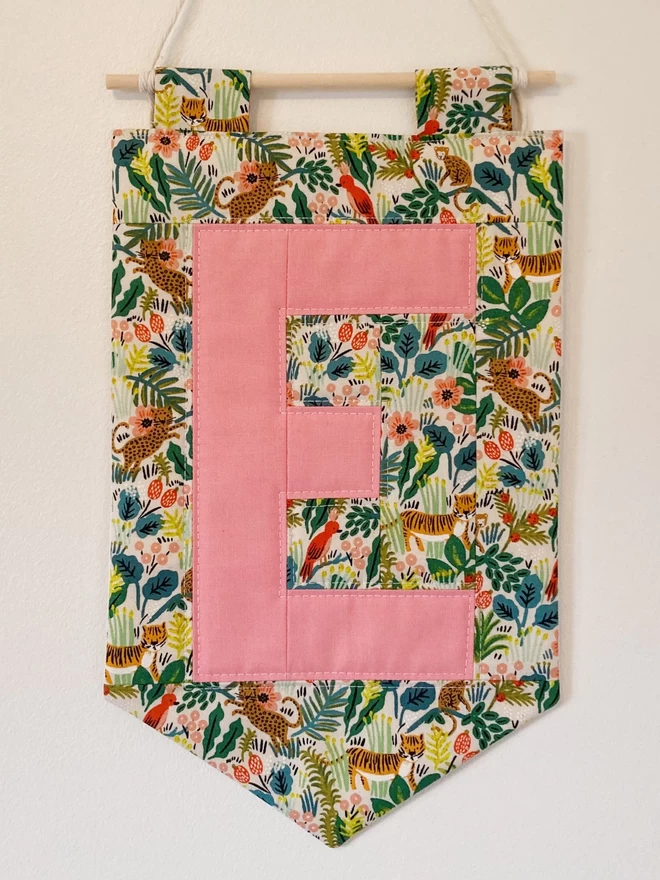 Cooper & Fred personalised quilted wall-hanging seen with a jungle style fabric and a pink letter 'E.'