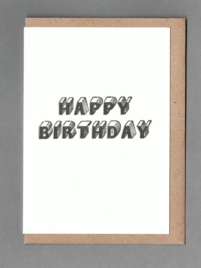 White card with black text reading 'Happy Birthday' with a brown envelope behind it