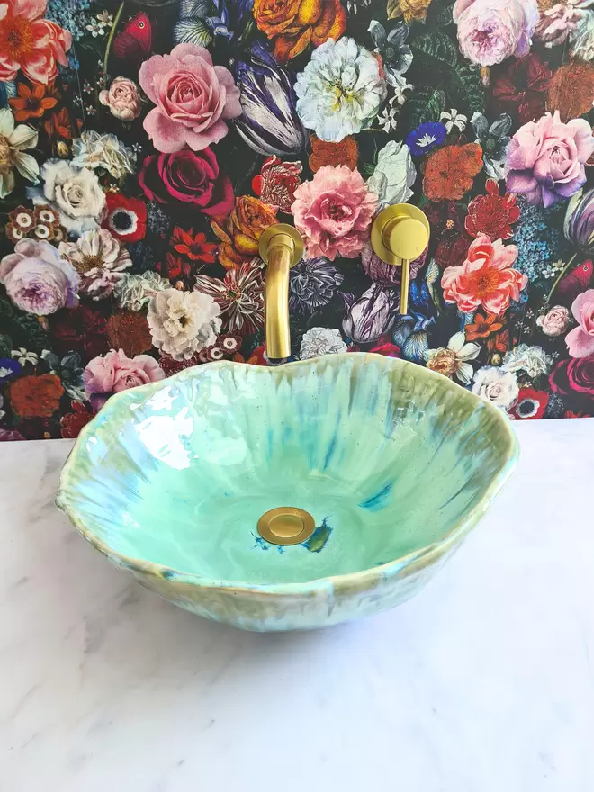 Handcrafted ceramic bathroom basin in a turquoise green blue glaze, hand-crafted sink, pottery basin, wc, bathroom, ensuite, modern bathroom, photographed against colourful floral wallpaper with gold taps, homeware, interiors, angled top front view
