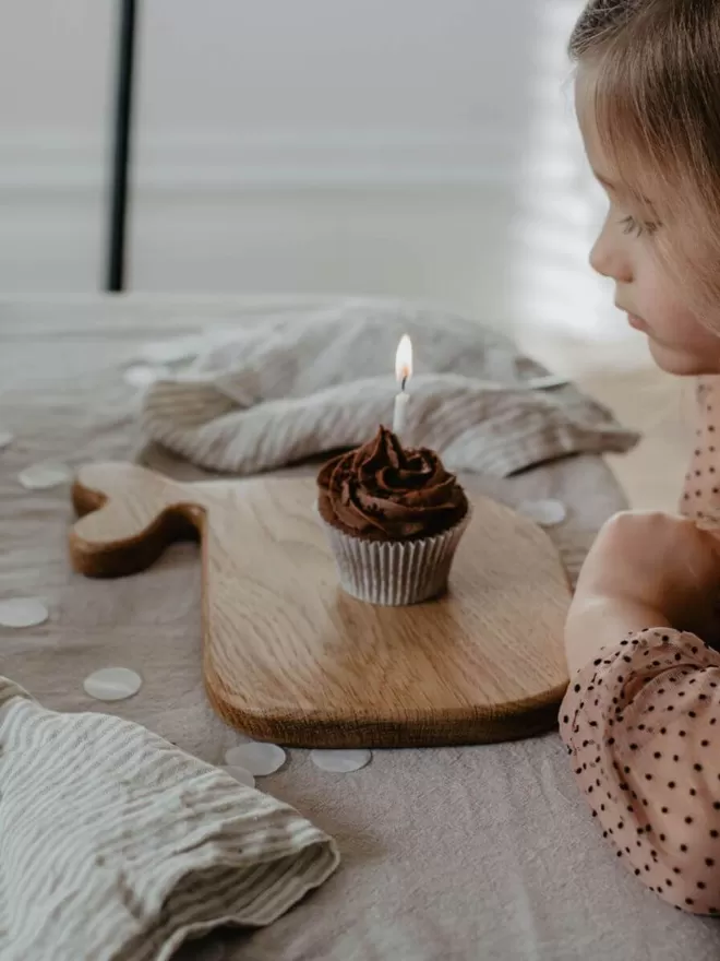 side view of a chocolate cupcake with a lit candle resting on a whale wooden serving board with a little girl ready to blow the candle out 