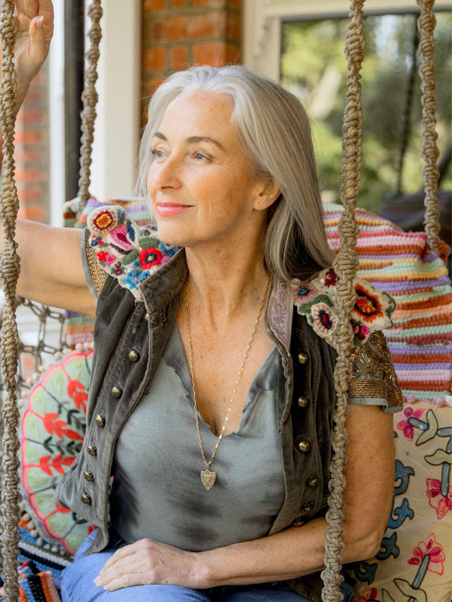 Woman with grey hair sitting in colourful chair wearing a gold paperclip chain necklace with a gold Joan of Arc shield charm necklace