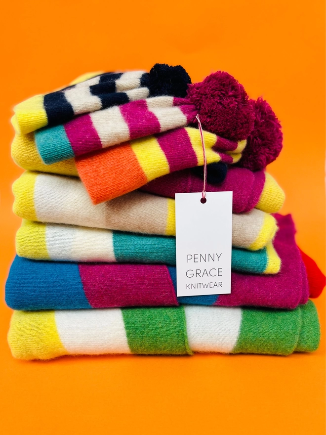 A stack of knitted blankets scarves and beanie hats on an orange background