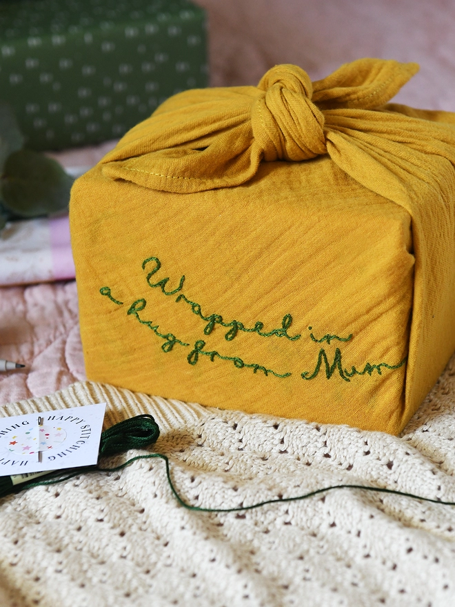 A gift wrapped in a mustard yellow cotton fabric wrap that has been embroidered with a special message. 