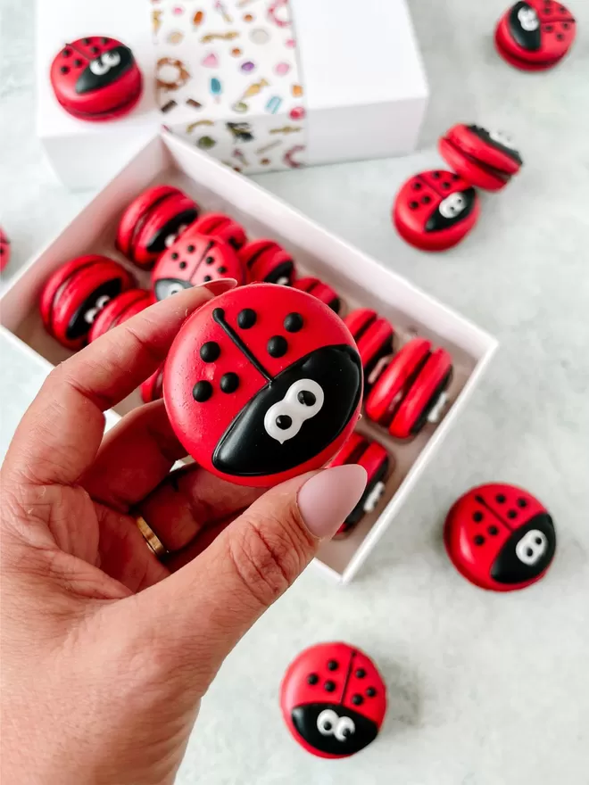 a person holding a red ladybug macaron in in front of a box of red ladybug macarons