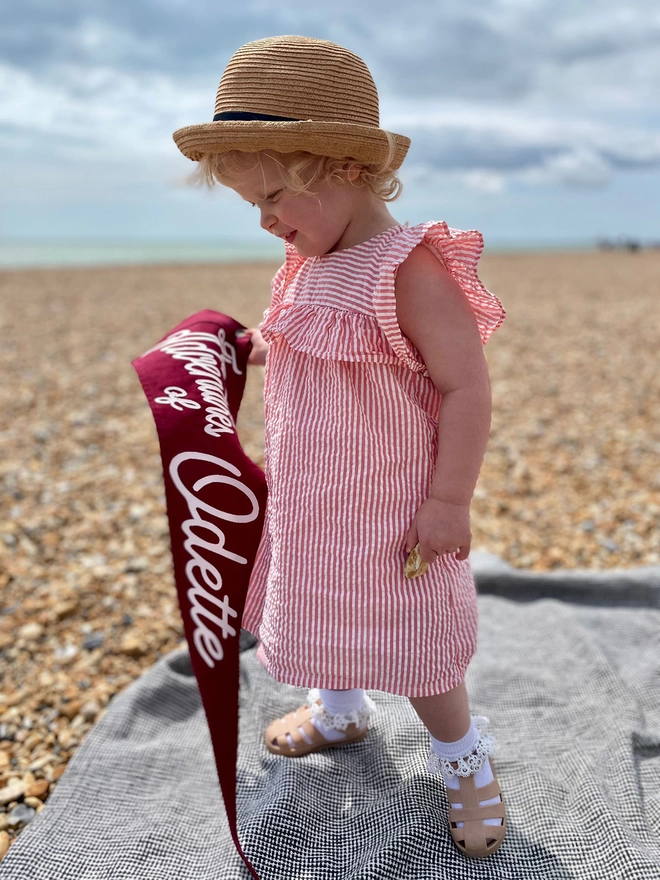 A little girl on a beach holding a pennant flag saying The Adventures of Odette on.