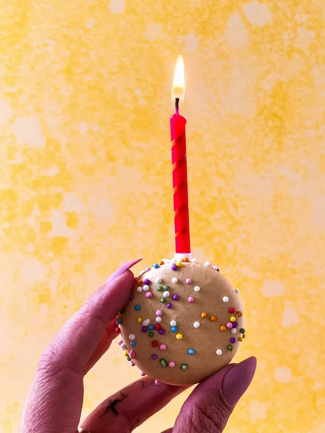 a hand holding a birthday cake macaron with a with a lit candle