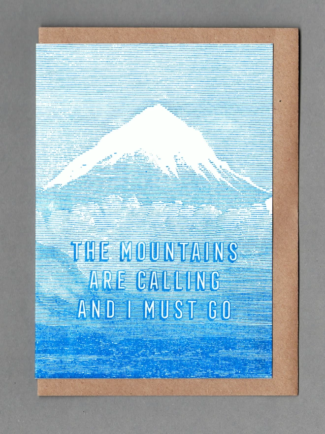 Blue and white card with a mountain on it and text reading 'THE MOUNTAINS ARE CALLING AND I MUST GO' with brown envelope behind it