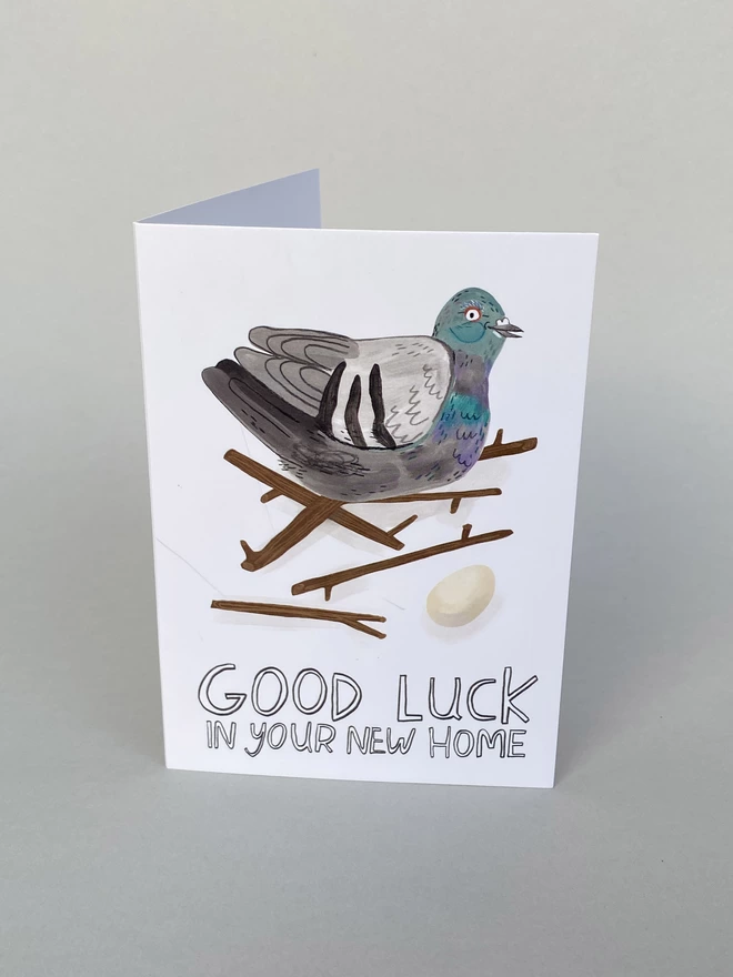 Illustrated Pigeon on terrible nest with an egg on a greeting card that reads Good luck in your new home