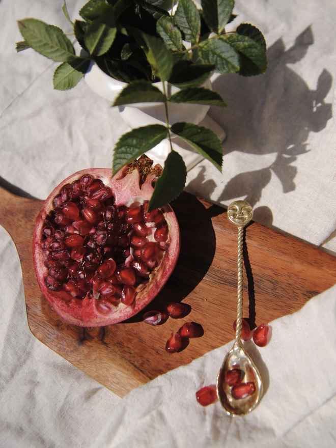 The image is of a gold toned brass spoon with a twisted handle and a hand carved moon face laying on a rustic wooden chopping board, next to half a pomegranate, some rose leaves and some cream coloured linen fabric 