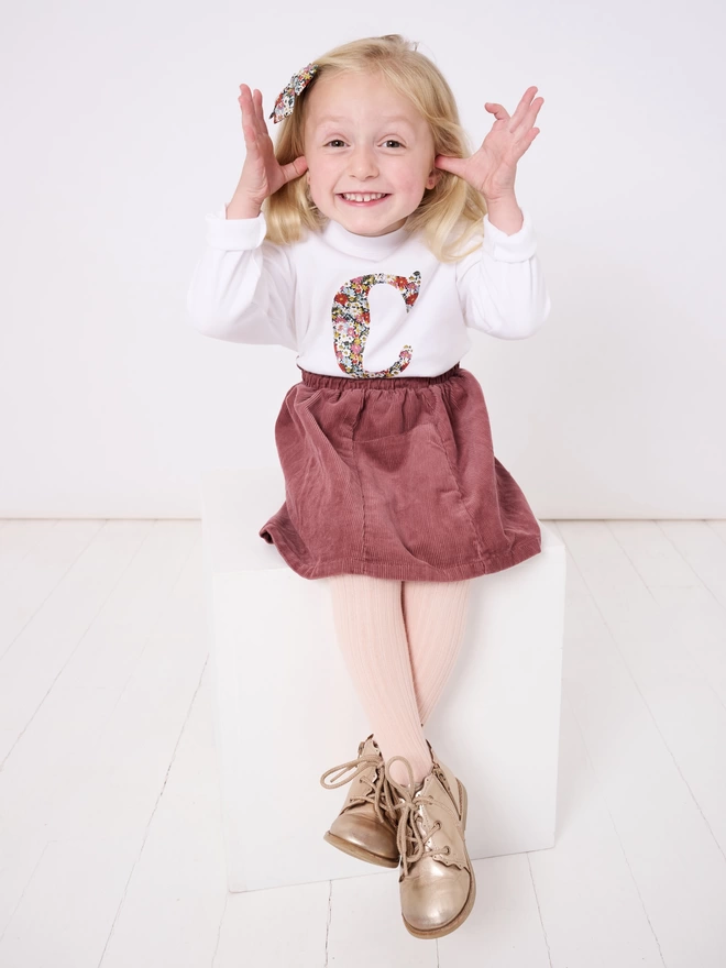 A white cotton long sleeve t-shirt appliquéd with an initial in a floral Liberty print, worn by a 4 year old girl 