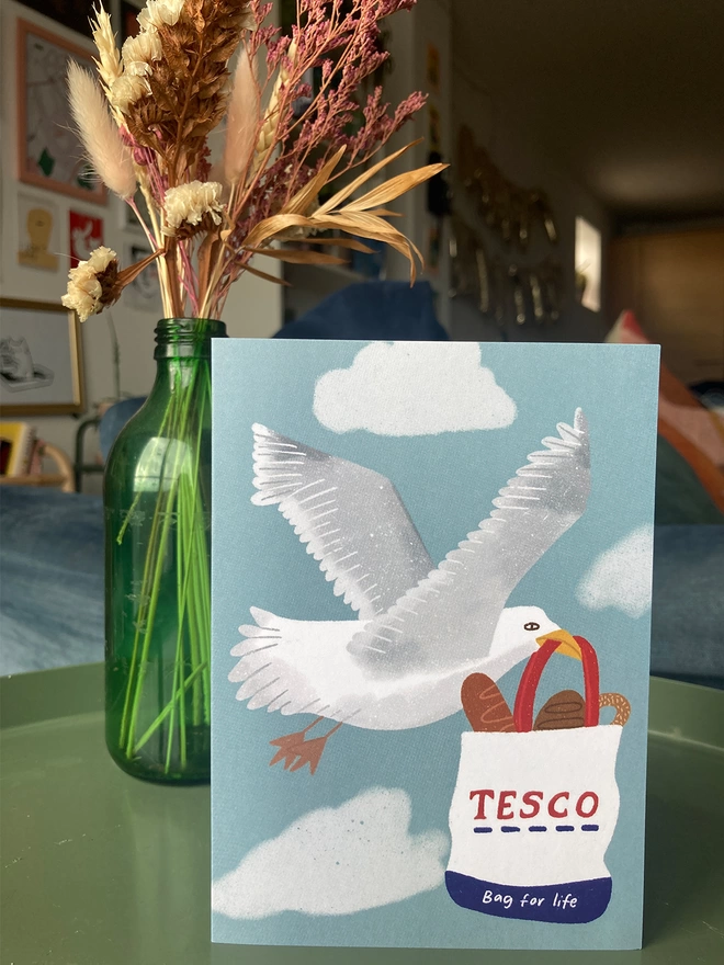 Greetings card illustrated with a seagull flying carrying a bag for life full of bread.