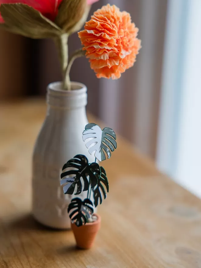 A miniature replica Variegated Monstera Deliciosa Albo paper plant ornament in a terracotta pot sat on a desk with a real climbing plant in the background