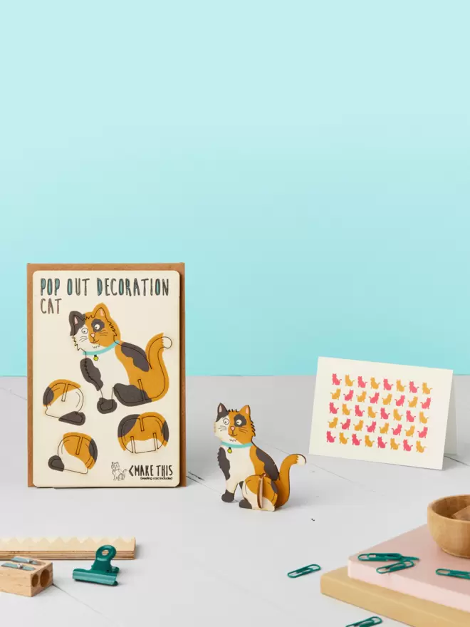 3D calico cat decoration and cat pattern greeting card and brown kraft envelope on top of a grey desk in front of a light blue coloured background