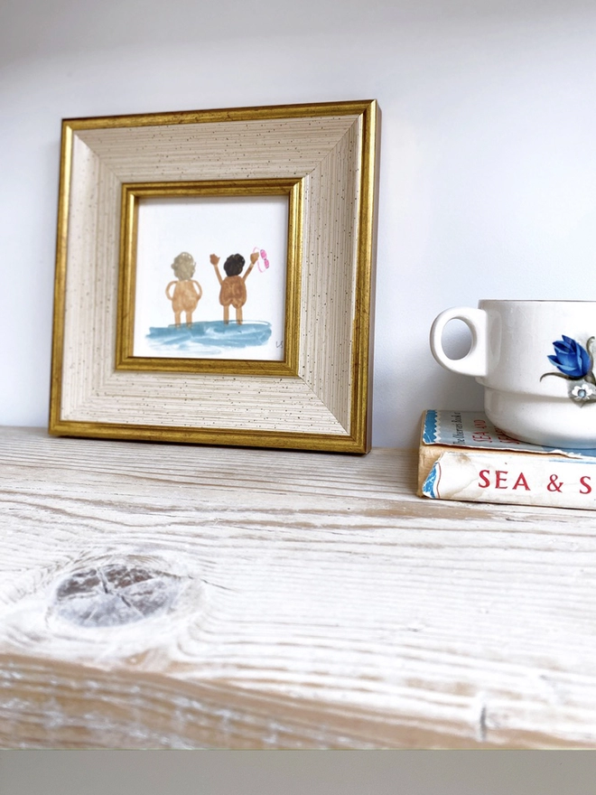 wooden shelf with vintage cup and framed picture of two naked people cheerfully looking out to sea with pink goggles