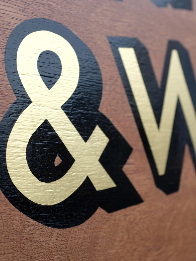 Close up of gold leaf ampersand outlined in black on dark stained wood. 