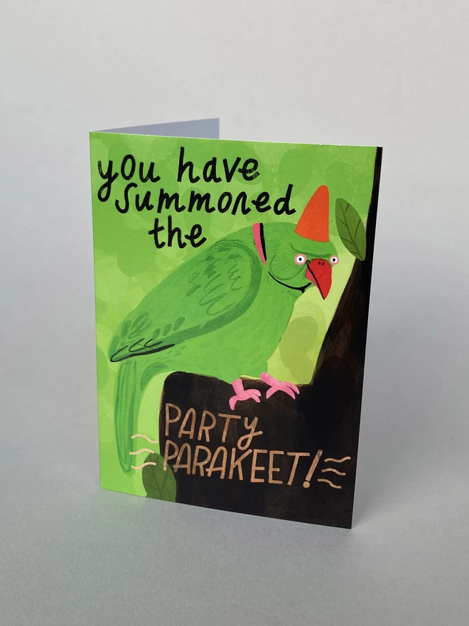 Illustration of a Green Parakeet on a tree in a red  party hat that reads you have summoned the party parakeet on an A6 greeting card