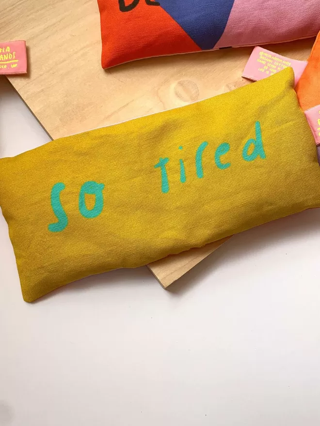Lavender eye pillow in yellow fabric with neon turquoise letters which read 'so tired' 