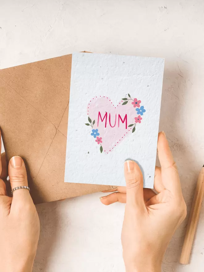Two hands holding a brown envelope and seeded paper card featuring a pink heart with flowers with 'Mum' in the centre.
