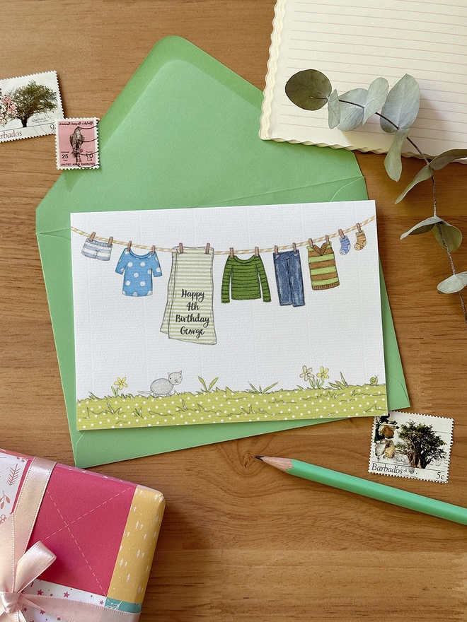 A personalised birthday greetings card with an illustrated washing line of children's clothes lays on a green envelope on a wooden desk.
