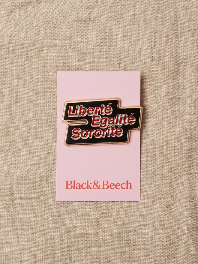 A black enamel pin with a gold edge with the words Liberté Egalité Sororité written in red with a white boarder, on a pink backing card 