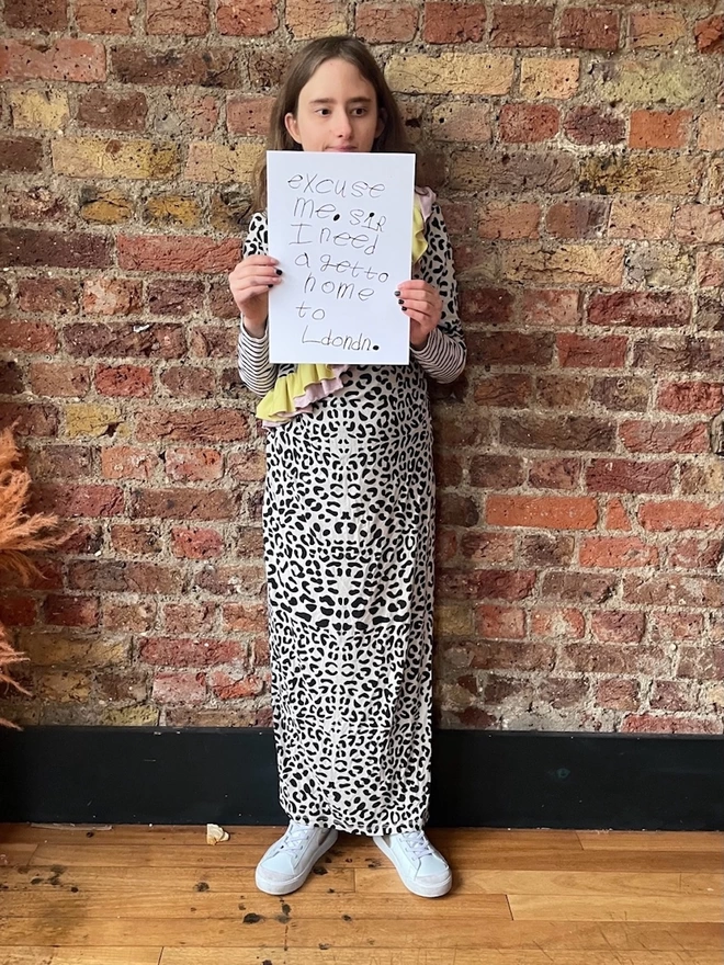 Piper a deaf, autistic teenage artist from hackney dressed in a full length leopard print dress hold hers artwork, black handwritten unique font on a white background in front of a brick wall.