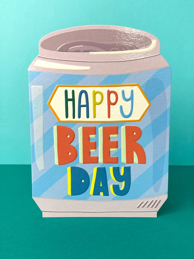 A vibrant die cut birthday card in the shape of a blue striped beer can with a rainbow coloured ‘Happy Beer Day’ message