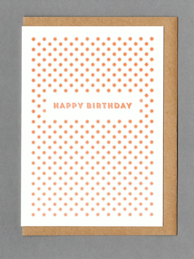White card with orange stars and orange text reading 'Happy Birthday' with a kraft envelope behind it