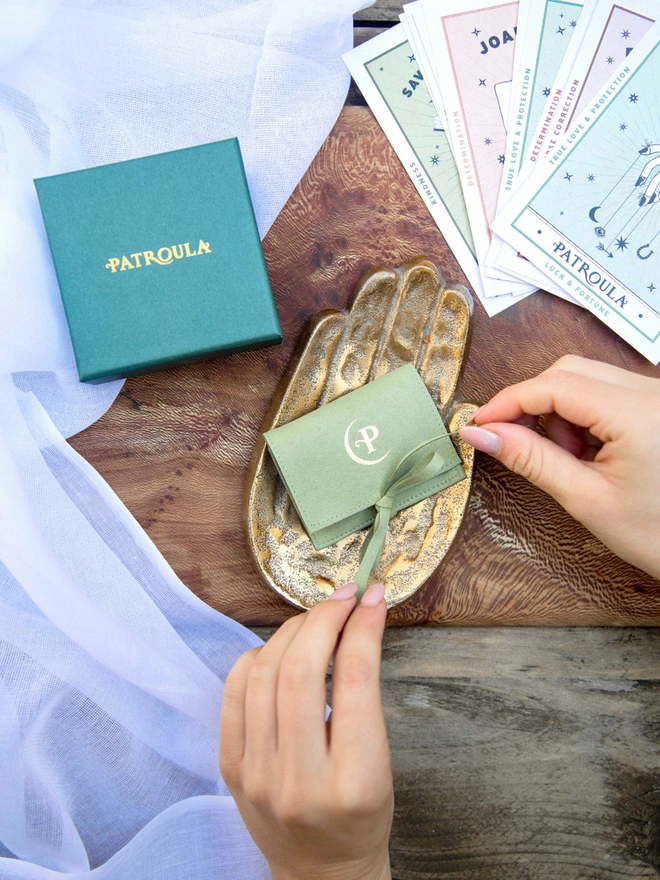 Hands tying a pale green jewellery pouch on a wooden table with a dark green patroula jewellery box in the background