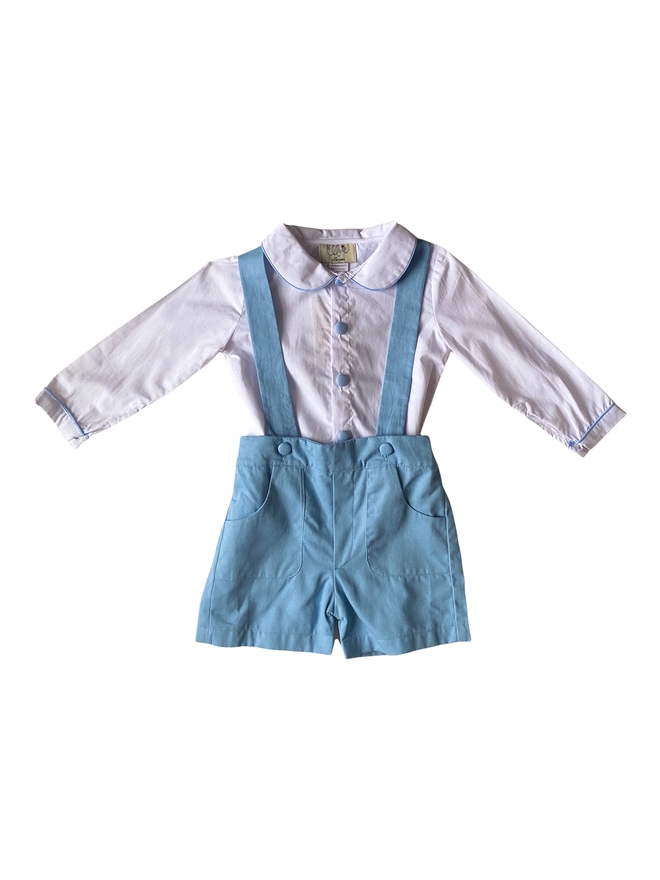 A cutout of a white shirt with a peter pan collar and blue piping and buttons styled with blue shorts with braces
