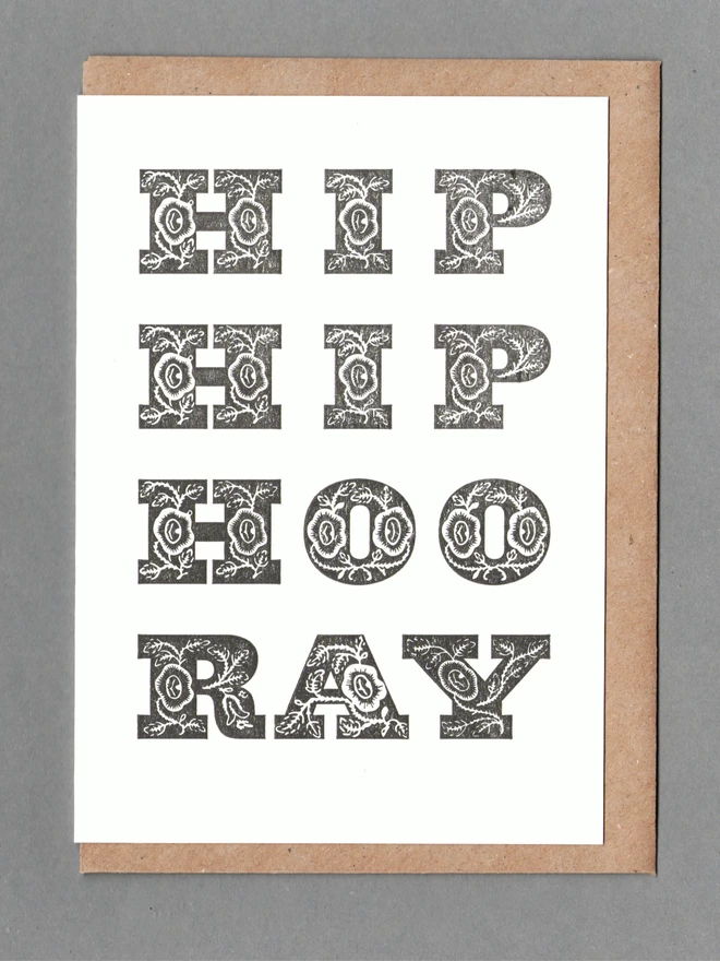 White card with chunky black text reading 'HIP HIP HOORAY' with a brown envelope behind it