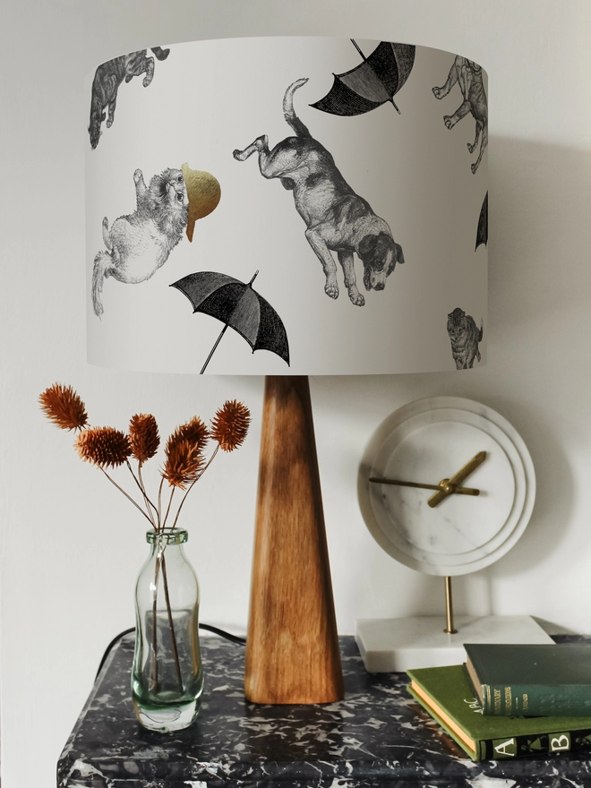 Drum Lampshade featuring cats and dogs ‘raining’ from the sky on a wooden base on a shelf with books and ornaments