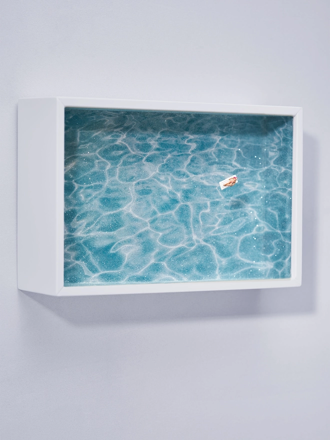 Miniature scene in an artbox showing a tiny naked sunbathing figurine on a lilo, floating on the glass on top of an artbox which looks like a sparkling turquoise swimming pool