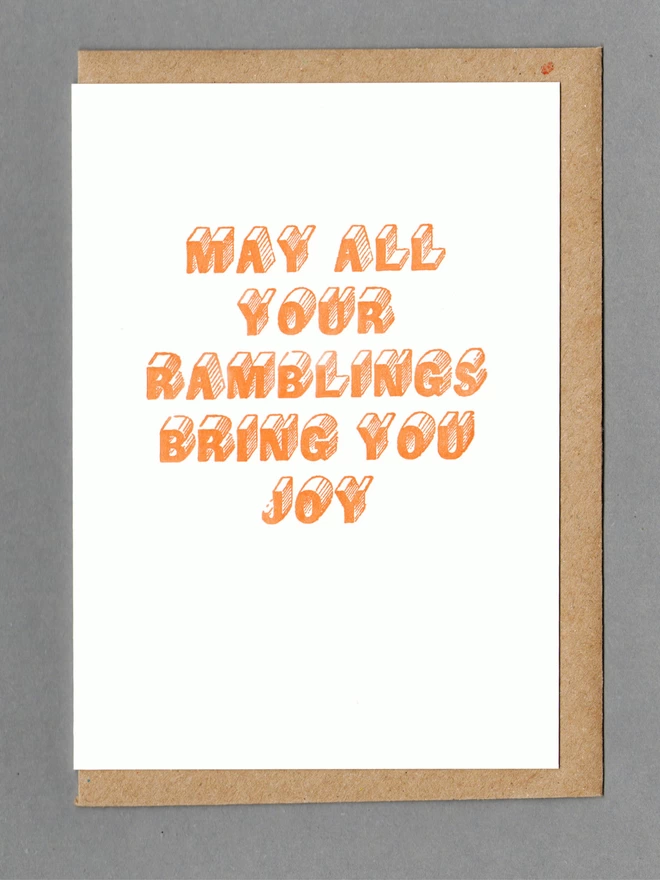 White card with orange text reading 'MAY ALL YOUR RAMBLINGS BRING YOU JOY' with a kraft envelope behind it