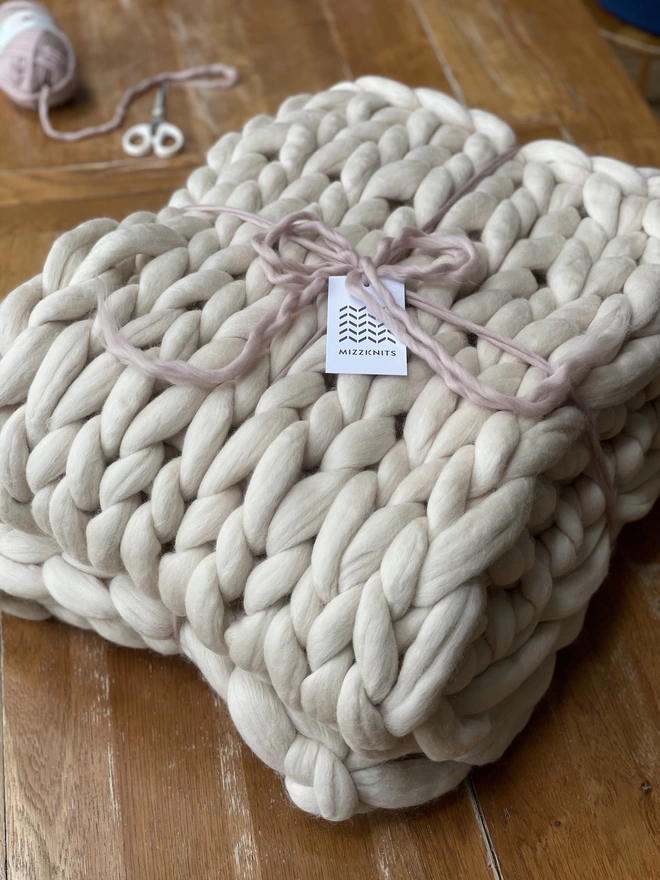 An oyster cream merino arm knitted blanket is folded and tied with a light pink yarn, with a mizzknits label, sitting on top of an oak table