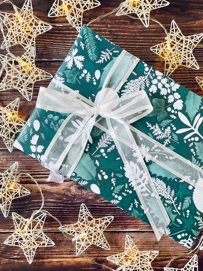 Present wrapped in recycled gift wrap in Christmas Winter leaf design, white ribbon, surrounded by star lights on wood background