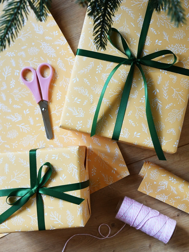 Several gifts wrapped in yellow botanical wrapping paper with green ribbons are on a wooden floor below a Christmas tree.