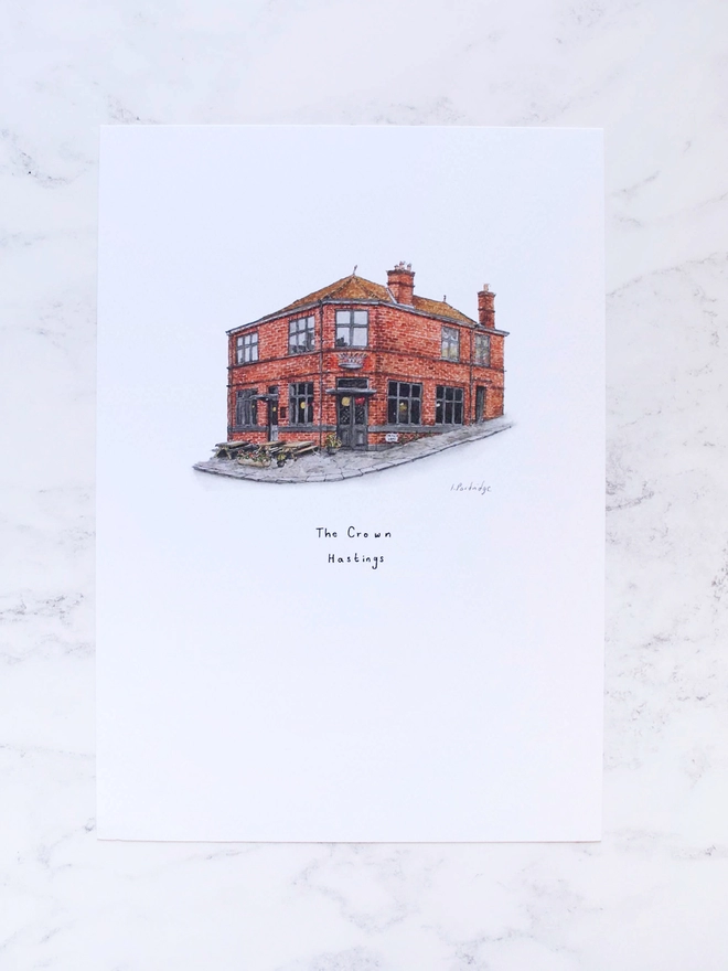 Watercolour illustration of The Crown in Hastings a beautiful brick fronted pub with a characterful appearance and a metalwork crown sitting above the entrance door. The illustration is a small watercolour painting in the centre of the white page, the page is laid out on a pale white marble background. 