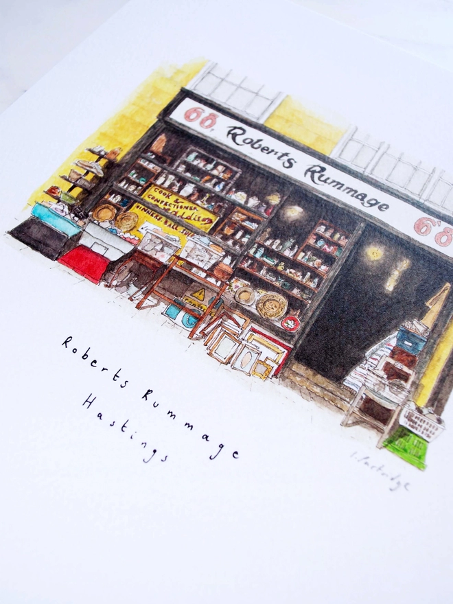 Watercolour illustration of Robert’s Rummage antique shop in Hastings, a black shopfront on a yellow building, the shop window is packed and antiques and bric a brac spill out into the street infront of the shop. The watercolour painting picks out tiny details of the scene and sits on a white piece of paper with the name written below. The photo is at an angle showing some of the intricate detail of the painting. 