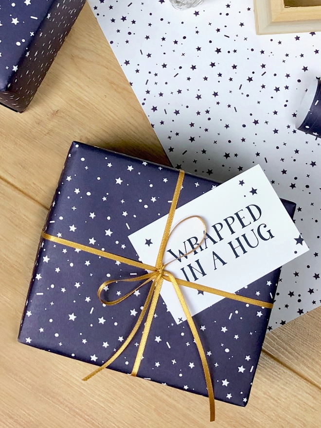 A gift wrapped in navy blue wrapping paper with a tiny star design and a gift tag that reads "Wrapped in a hug" is on a wooden surface.