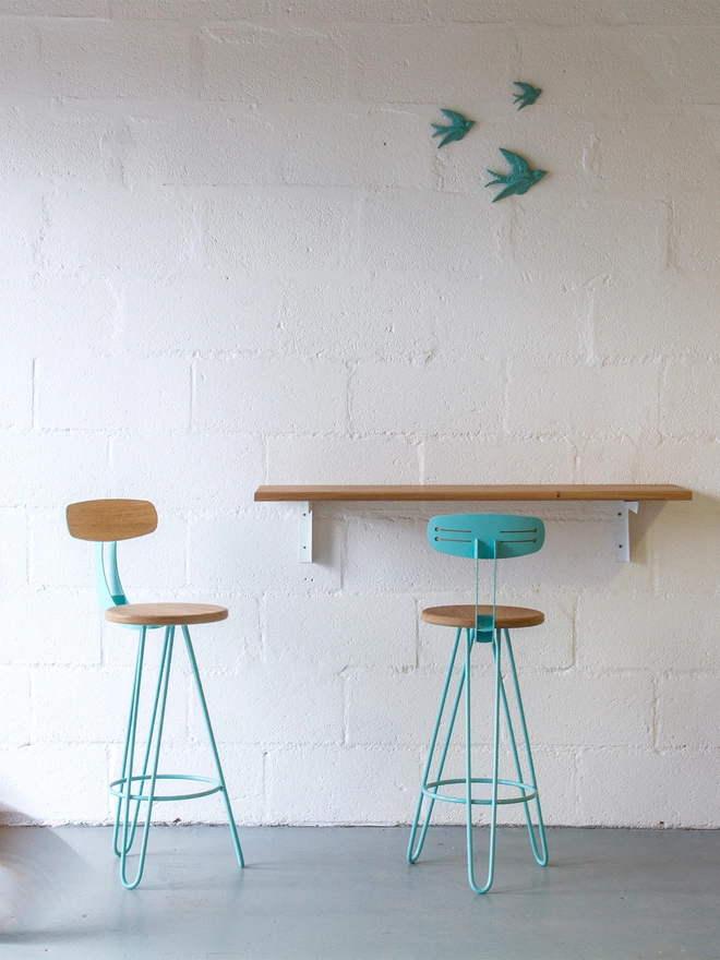 pair of hairpin leg bar chairs with turquoise legs and backs and oak seat and back rest, against a bar with a set of three ceramic turquoise birds hanging above