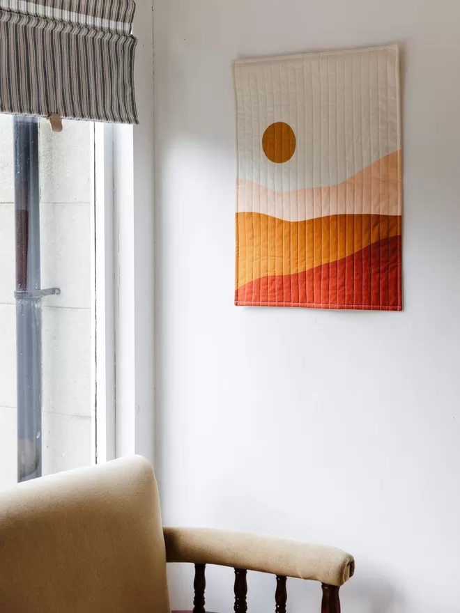 Earth Quilt Hanging On White Wall Above Cream Chair Next To Window